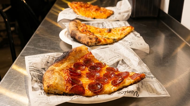 Three slices of pizza, including pepperoni and cheese, on newspaper-lined plates on a metal counter.