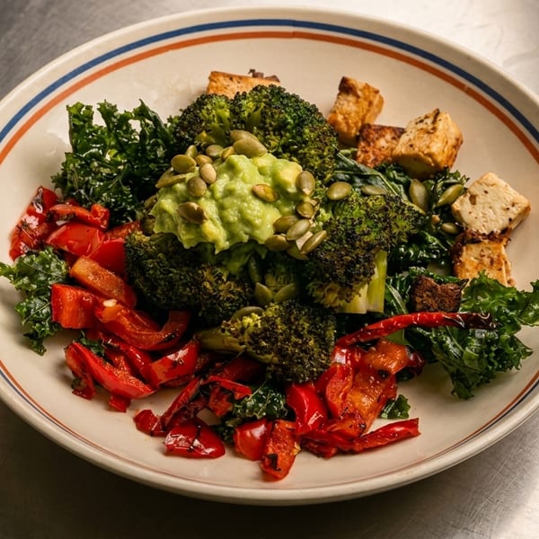 A colorful vegan bowl with broccoli, kale, red peppers, tofu, guacamole, and pumpkin seeds.