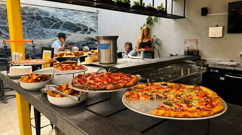 A buffet table with various pizzas and potato wedges, with people dining in the background.