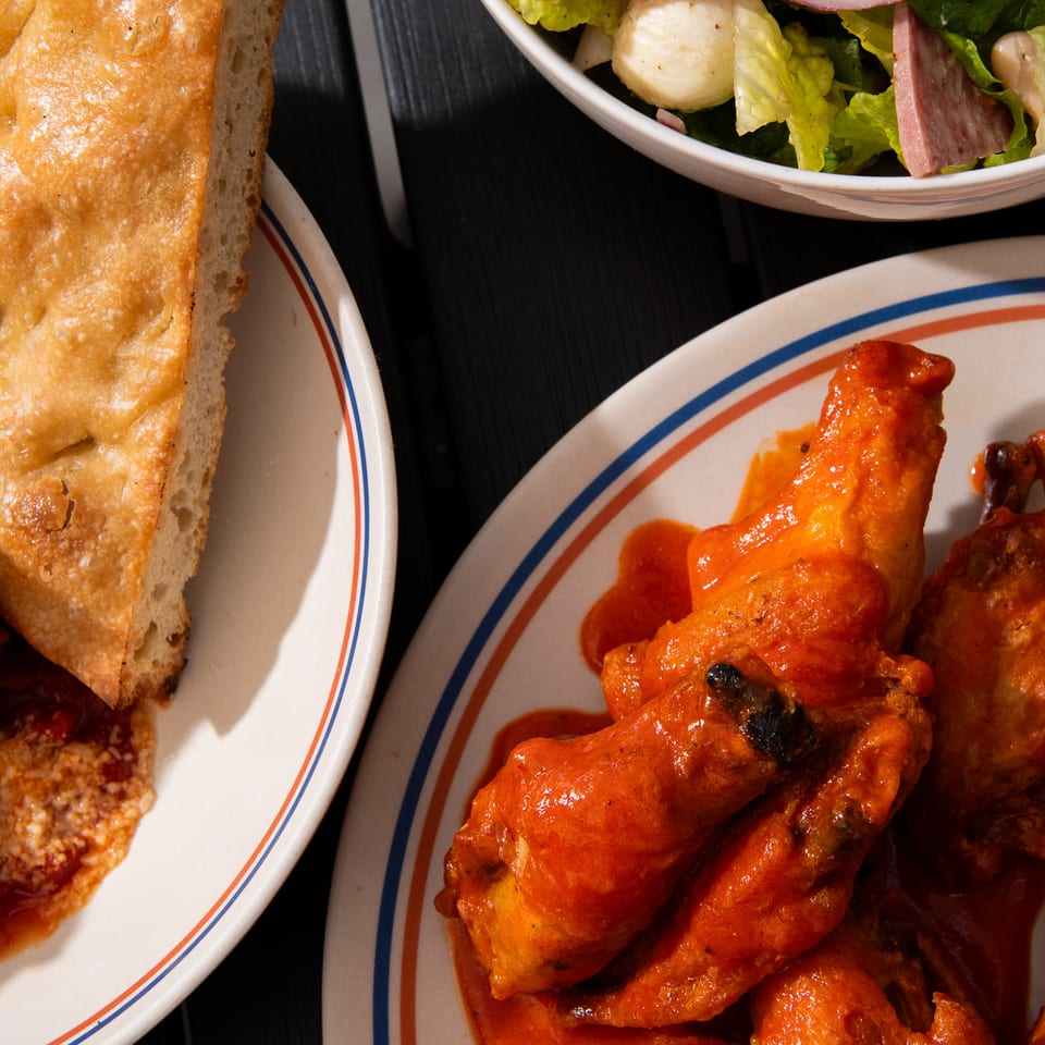 Buffalo wings with baguette, salad, and a partially visible sauced dish arranged on plates against a dark table.