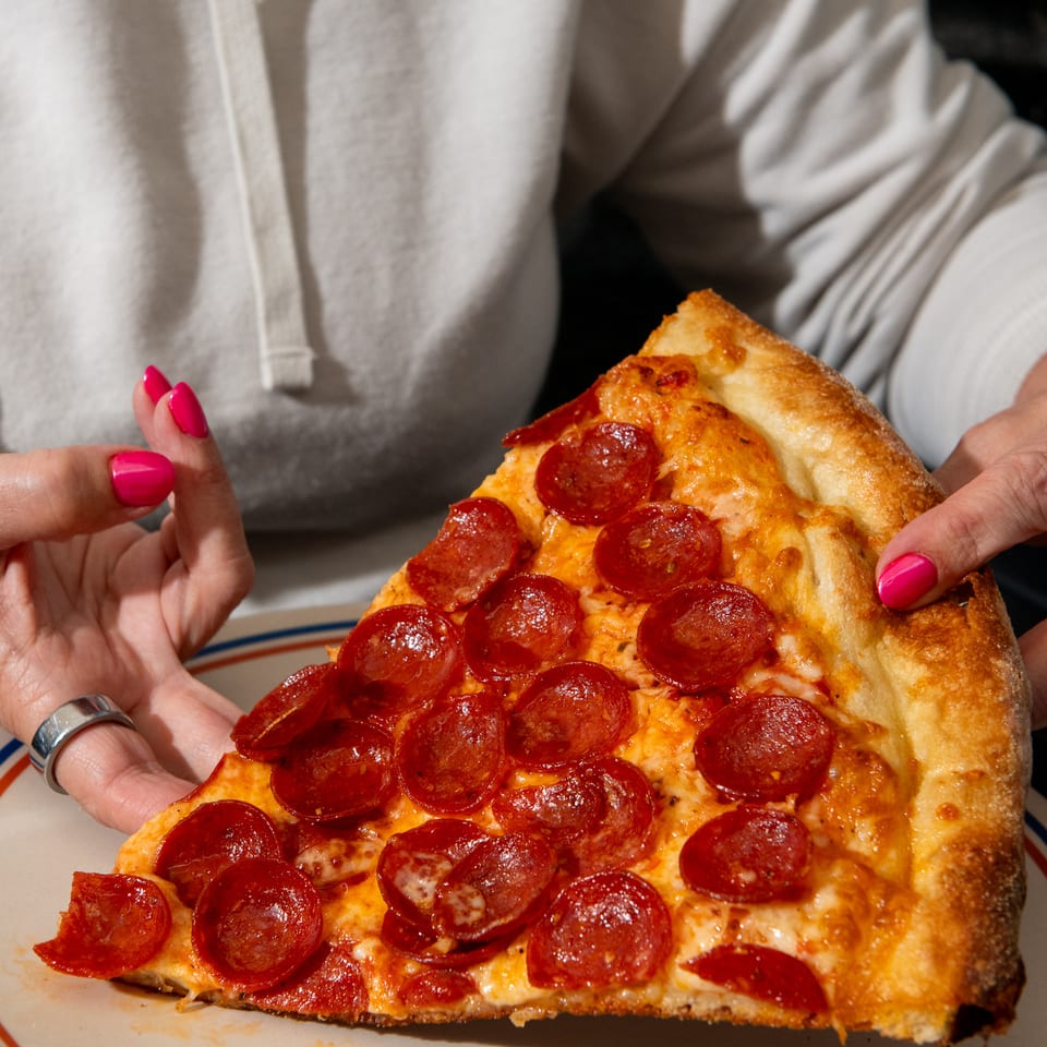 Person holding a large pepperoni pizza slice over a striped plate, with a golden crust and melted cheese.