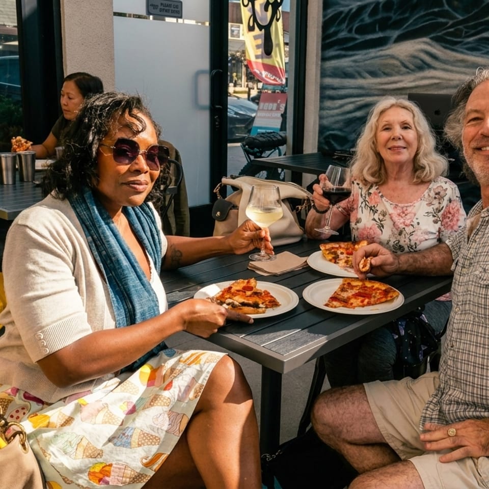Three friends enjoy pizza and drinks at an outdoor restaurant table with a mural behind them.
