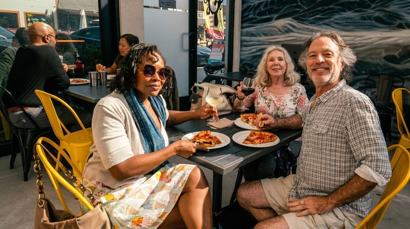 Three friends enjoy pizza and drinks at an outdoor restaurant table with a mural behind them.